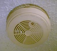 A white, round ceiling-mounted smoke detector with a circular vented grille, fixed to a ceiling