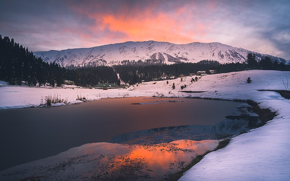 A snow-covered Himalayan mountain valley at sunset, with a partly frozen lake, snowy meadows and pine forest below the peaks.
