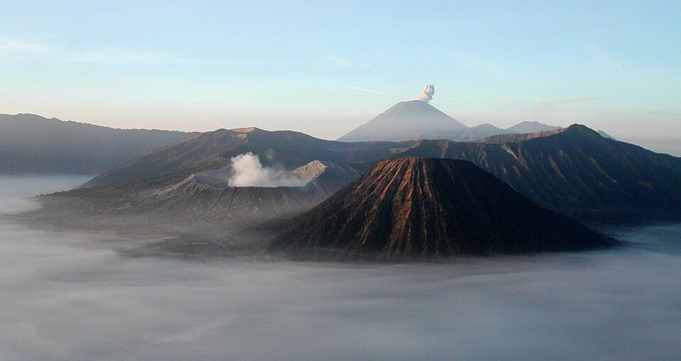 Dark volcanic cones rising above low morning mist, with a distant smoking peak against a pale sky (the Bromo-Tengger-Semeru volcanoes, East Java).
