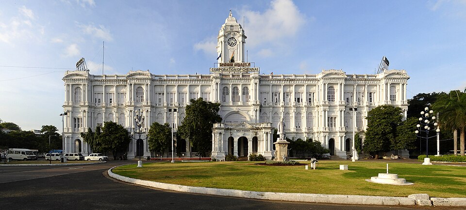 A large white colonial-era building with a central clock tower, fronted by lawns, bearing a 'Corporation of Chennai' banner (the Ripon Building).