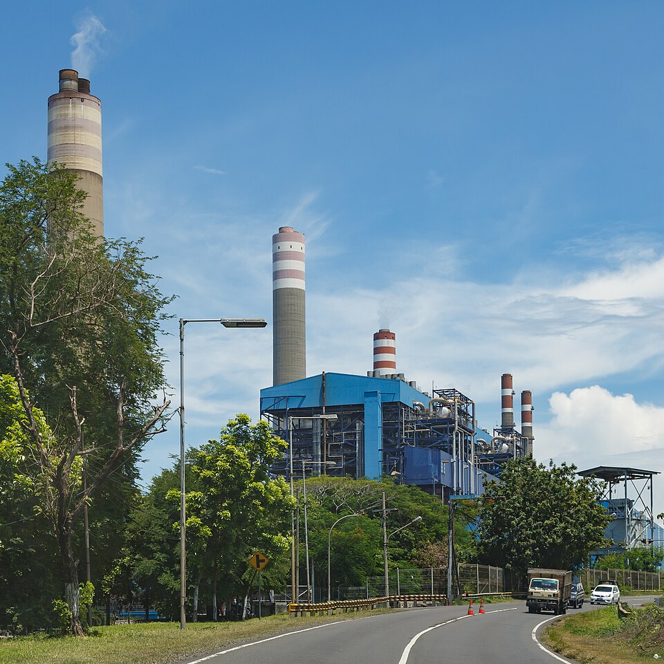 A large coal-fired thermal power plant with several tall concrete chimneys, one emitting smoke, and blue industrial buildings beside a road.