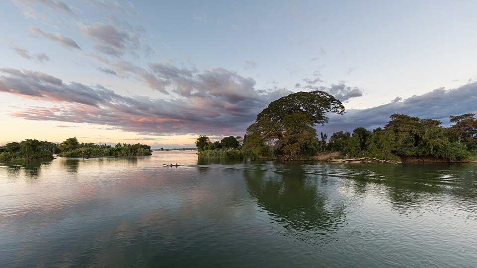 A broad calm river at dusk with forested islets, a small boat, and pink-tinged clouds reflected in the water (the Mekong).