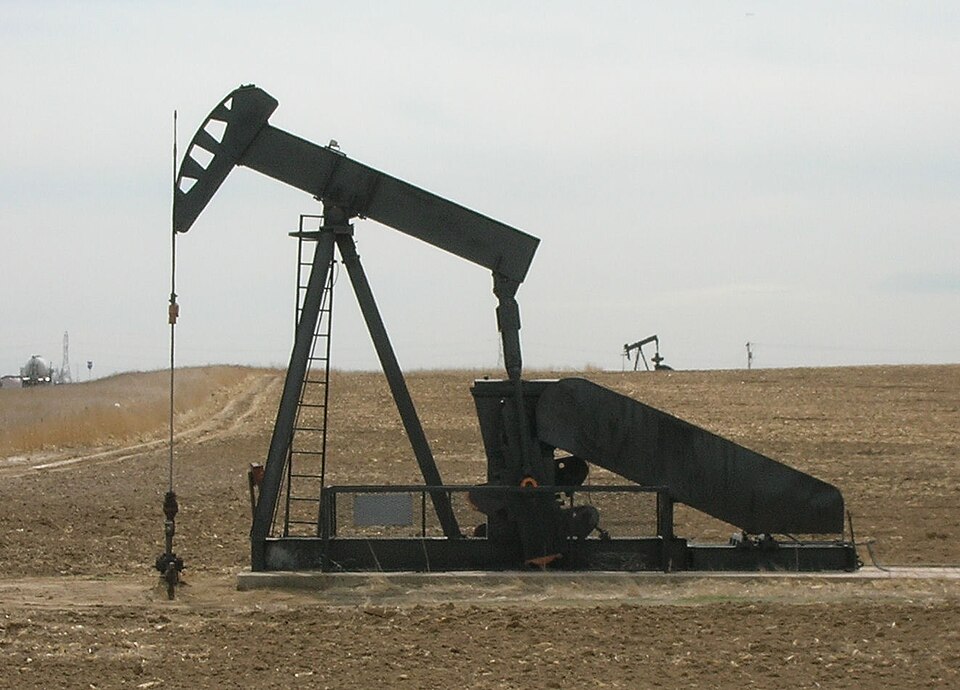 A black oil-well pump jack (nodding donkey) standing in a bare field, with a second pump jack in the distance.