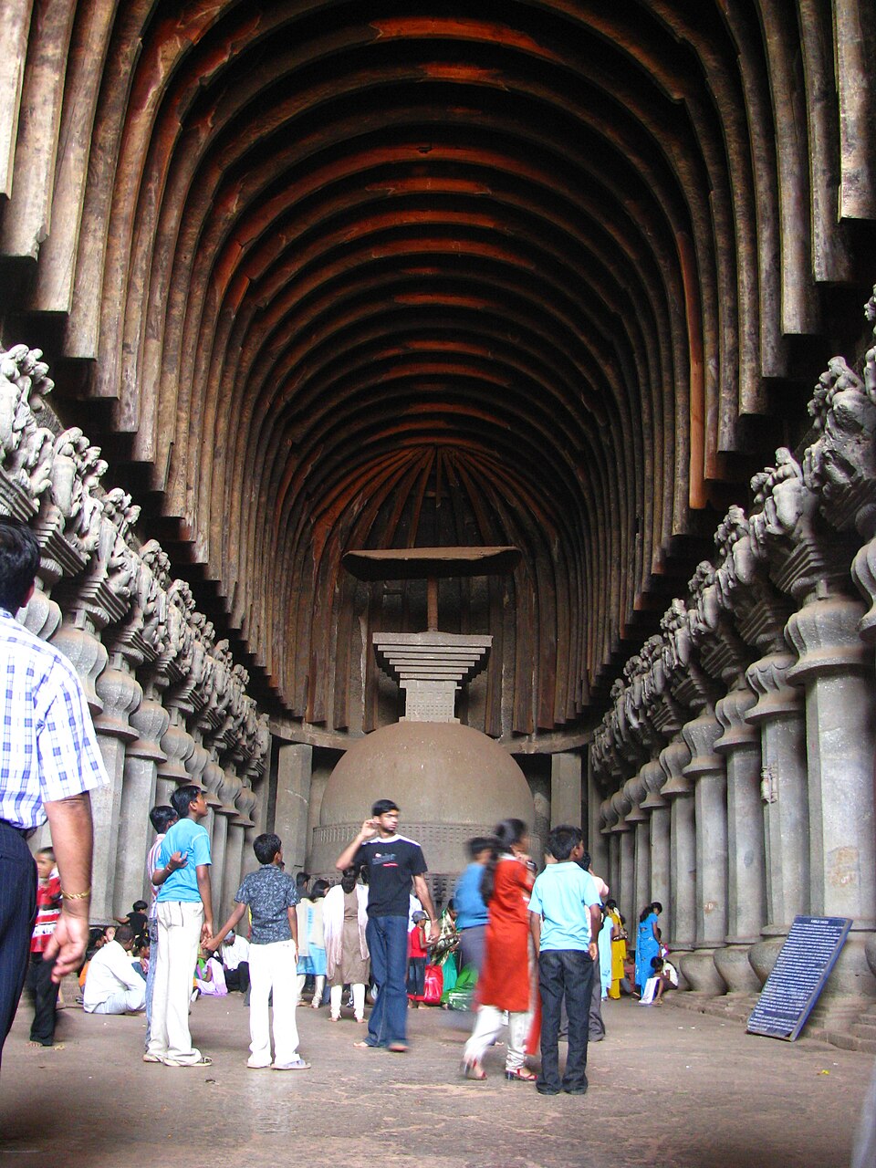 The vaulted interior of the rock-cut Karla chaitya hall, with rows of carved stone pillars leading to a domed stupa while visitors walk on the floor.