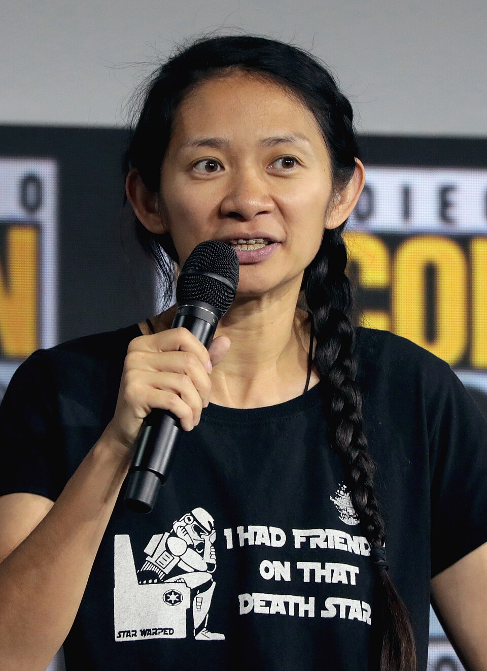Film director Chloe Zhao speaking into a handheld microphone, wearing a black T-shirt with a long braid over one shoulder.