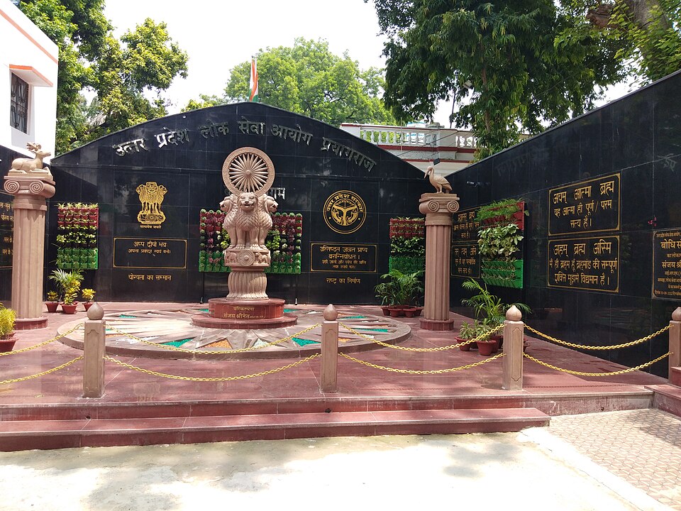 The Uttar Pradesh Public Service Commission campus in Prayagraj, showing a replica Ashoka Lion Capital and a black granite wall inscribed with the Commission's name in Hindi.
