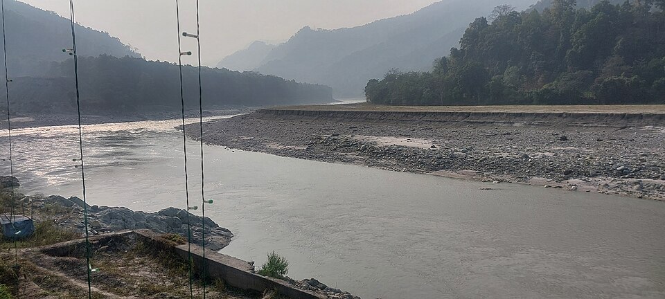 The Teesta River flowing between forested Himalayan hills near Rangpo in Sikkim, with a broad stony bank and a misty valley behind.