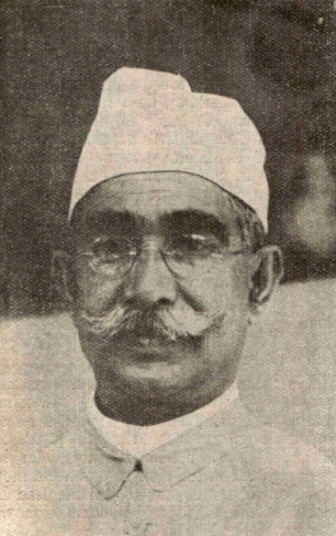 A black-and-white portrait of Mukhtar Ahmad Ansari, an elderly man wearing a white cap, round spectacles and a moustache.