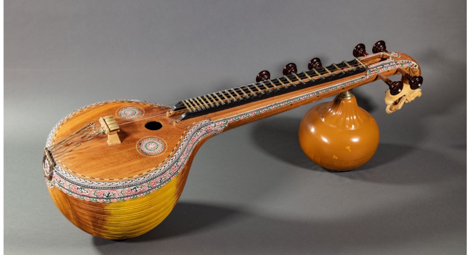 A Saraswati veena, a South Indian string instrument with a large rounded wooden body, a long fretted neck and a gourd resonator, on a plain grey background.