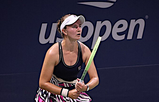 A female tennis player in a white cap and dark top holding a racket in a ready stance in front of a US Open backdrop.