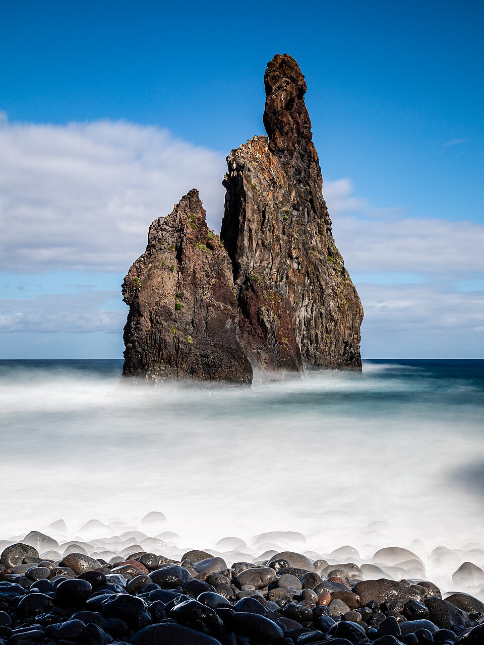A tall jagged volcanic sea stack rising out of the ocean beside a black-pebble beach under a blue sky, on the coast of Madeira.