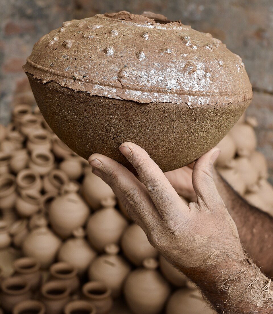 A person's hand holding up a large rounded brown clay earthen pot, with rows of stacked unfired earthen pots piled behind.