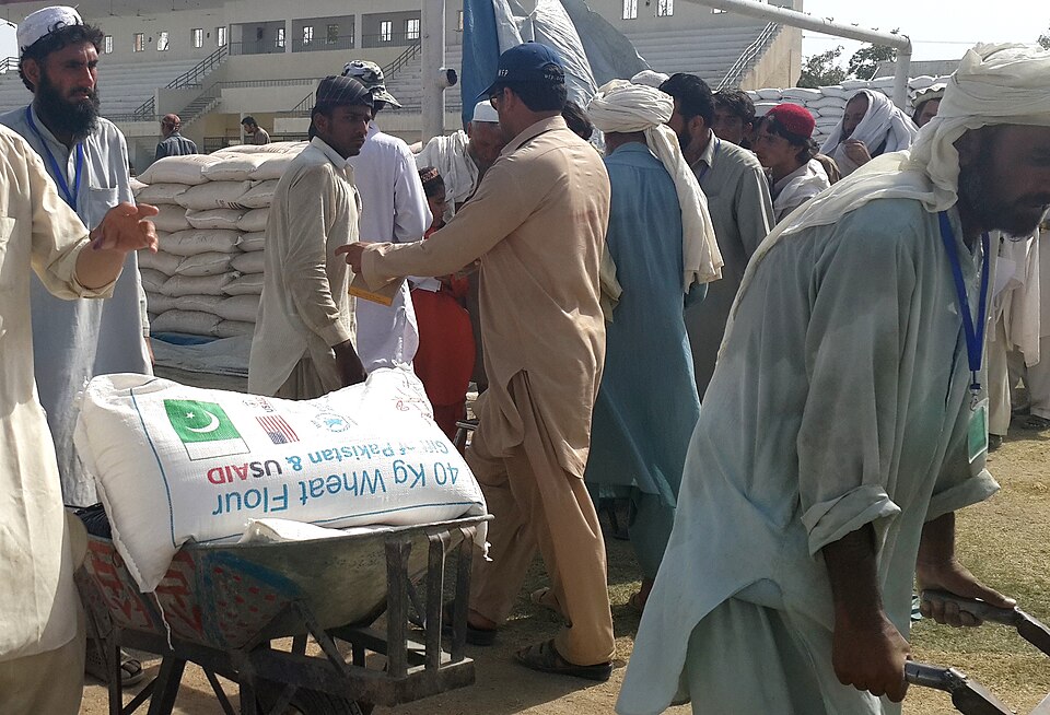 People queue at an open-air humanitarian food-distribution point where relief workers hand out large sacks of wheat flour, with more sacks stacked behind.