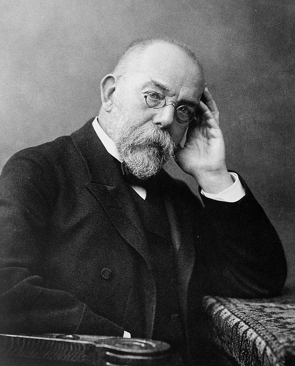A black-and-white studio portrait of an elderly bearded man wearing round pince-nez spectacles, resting his head on his hand (Robert Koch).