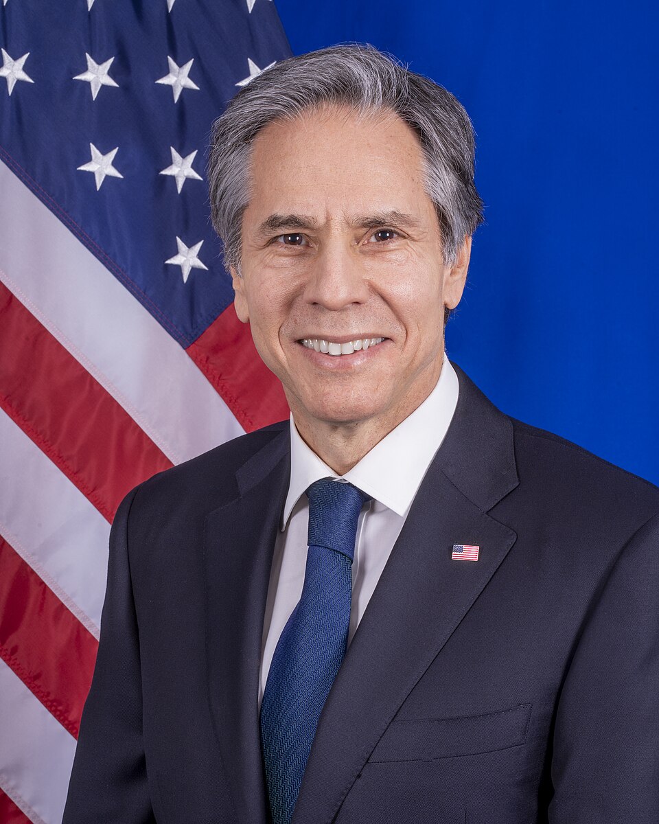An official portrait of a grey-haired man in a dark suit and blue tie smiling in front of the United States flag (Antony Blinken).