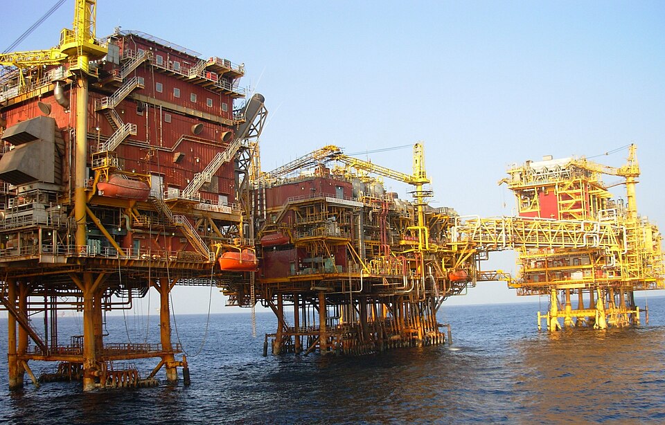 A large offshore oil-and-gas platform complex of red and yellow steel structures standing on legs above the sea, linked by bridges and fitted with cranes and lifeboats.