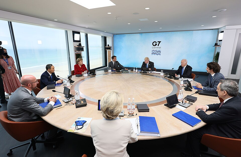 World leaders seated around a large round table beneath a 'G7 Cornwall' backdrop, with a beach visible through the windows, at the 2021 G7 summit.