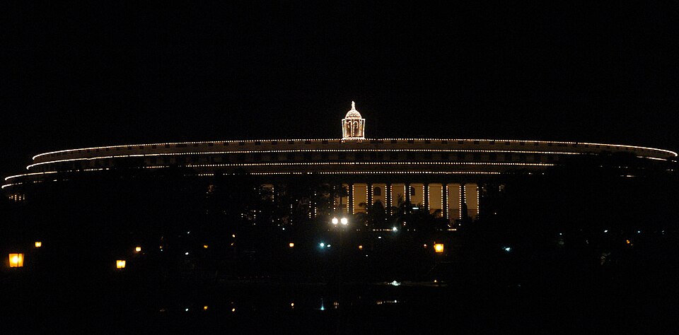 The circular Parliament House of India outlined in strings of lights at night, its central dome brightly illuminated against a dark sky.