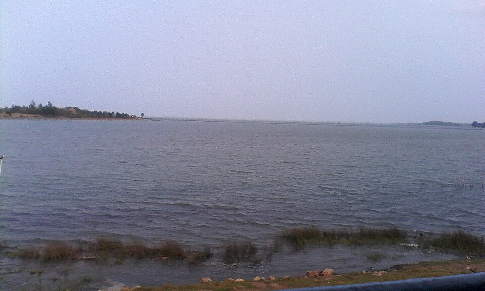 A broad expanse of open water at Chilika Lake in Odisha under a hazy sky, with a low distant shoreline and a grassy bank in the foreground.