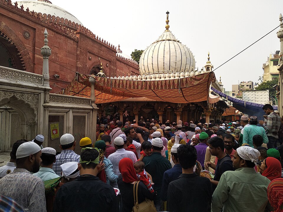 A dense crowd of pilgrims gathered before the white marble domed shrine of Hazrat Nizamuddin Auliya in Delhi, with ornate red-sandstone structures alongside.