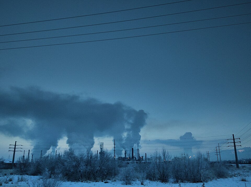 An industrial plant at dusk with numerous tall stacks releasing billowing plumes of smoke and steam into a dim blue sky above a snowy landscape crossed by power lines.