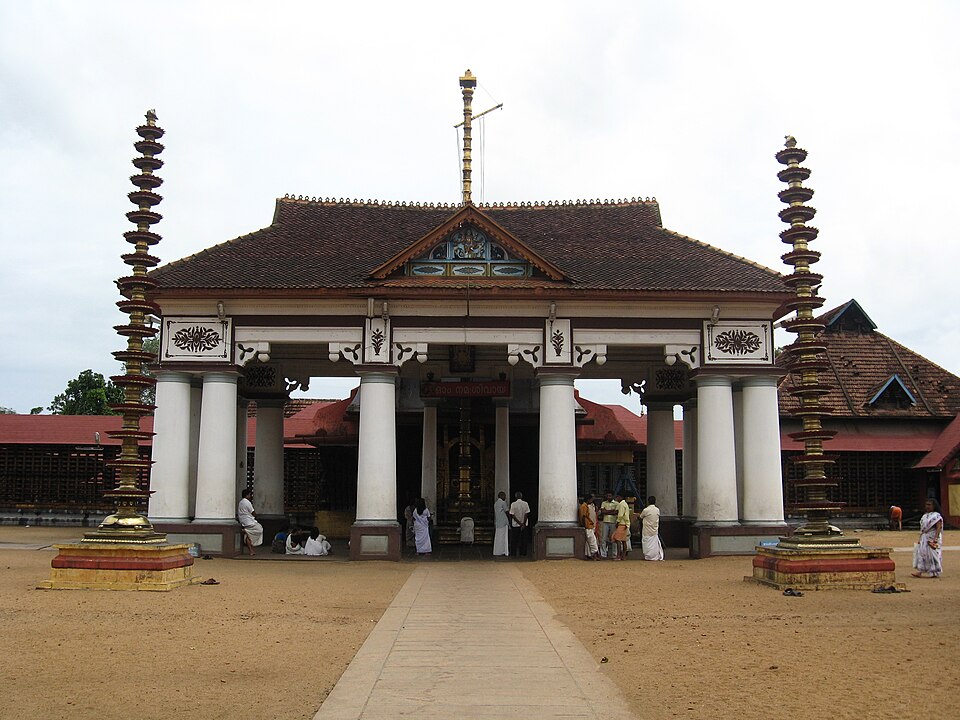 The gabled entrance of the Vaikom Mahadeva Temple in Kerala, flanked by two tall tiered brass lamp-columns, with devotees at the doorway and a sandy courtyard in front.