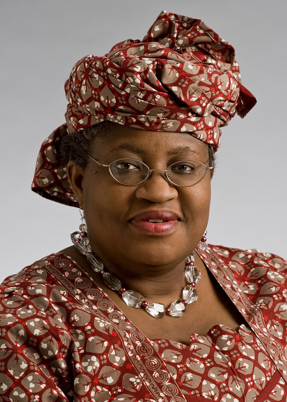Portrait of Ngozi Okonjo-Iweala, a woman in a red-and-tan patterned Nigerian dress and matching headwrap with glasses and a beaded necklace, smiling against a plain background.