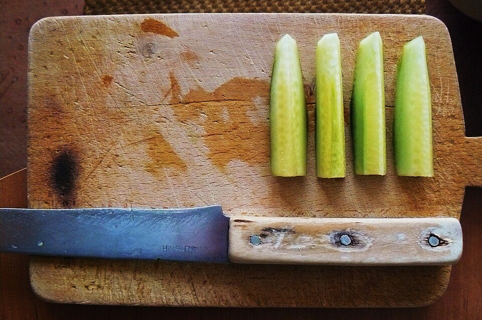 Four sticks of freshly cut green cucumber lined up on a worn wooden cutting board beside a kitchen knife.