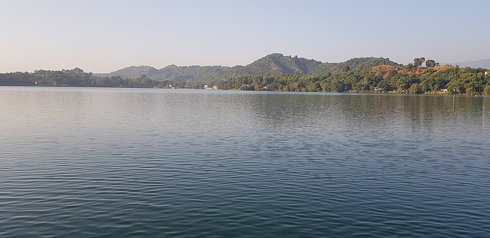 A calm, wide expanse of lake water with low forested hills and a clear sky in the background.