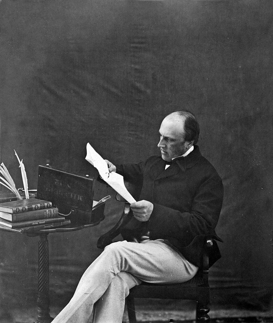 A black-and-white photograph of a seated 19th-century man in a dark coat reading a document at a small table stacked with books.