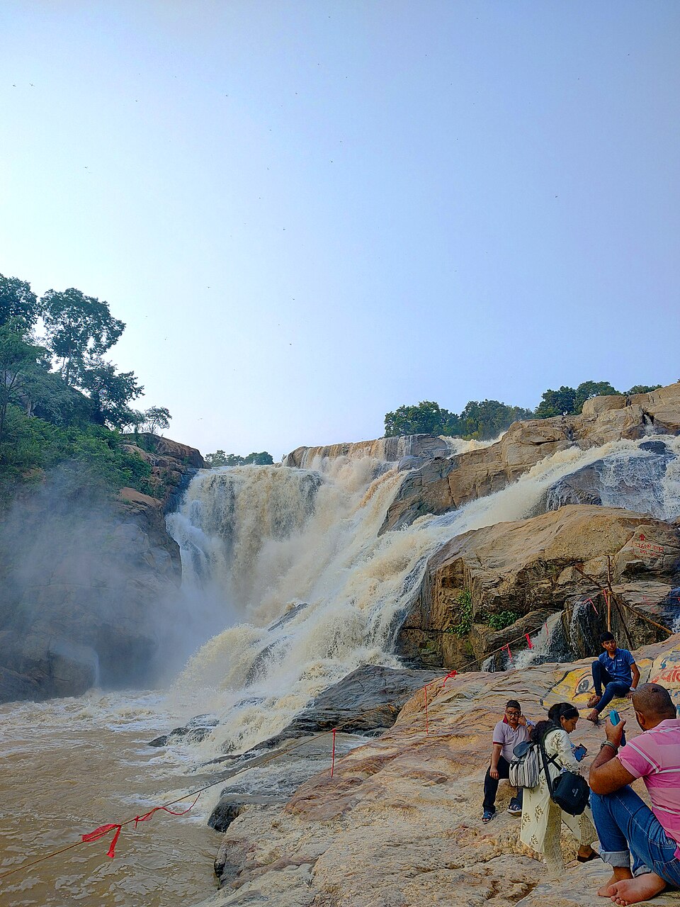 A wide, powerful waterfall plunging in white cascades over rock ledges into a river, with forested cliffs and visitors sitting on the rocks in the foreground.