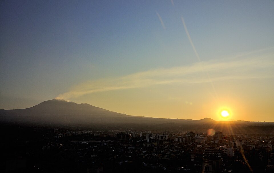 The dark silhouette of Mount Etna with a faint plume rising from its summit at dusk, above a city and a bright low sun.