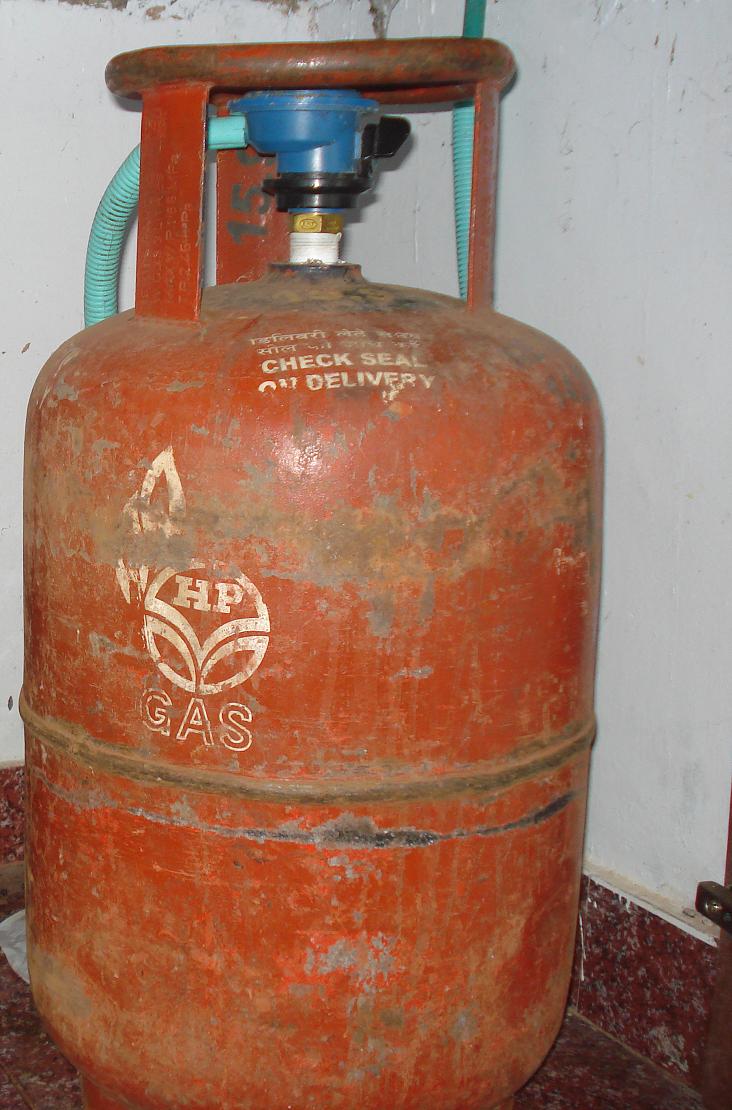 A worn orange domestic cooking-gas cylinder with a blue regulator and a green rubber hose, standing against a wall.