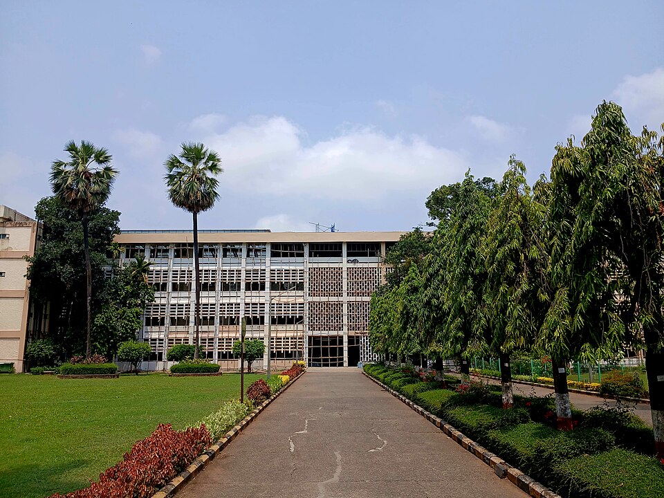 A long multi-storey institutional building seen at the end of a tree-lined pathway, with lawns, flower beds and palm trees in the foreground.