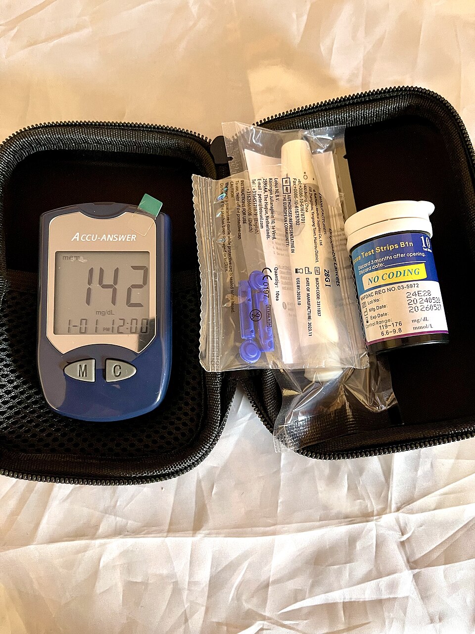 A blue handheld blood-glucose meter showing a reading of 142 mg/dL, beside a vial of test strips and a packet of lancets inside an open zippered case.