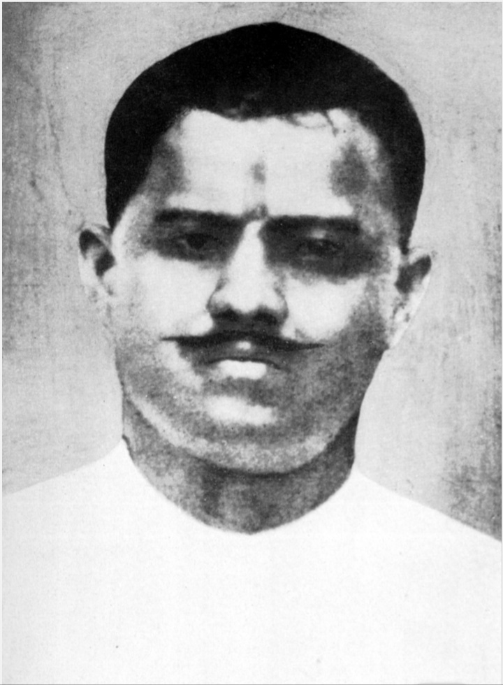 A black-and-white portrait photograph of a young man with a moustache and a forehead tilak, wearing a white garment.