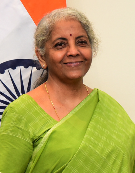 An official portrait of a grey-haired woman in a green sari standing in front of the Indian national flag.