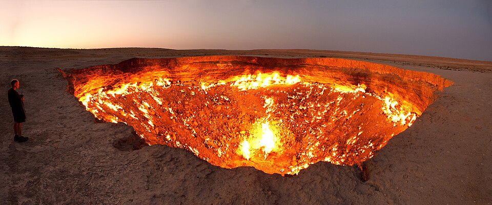 The burning Darvaza gas crater at dusk, flames covering the floor and walls of a wide pit in flat desert, with a small figure standing at the rim for scale