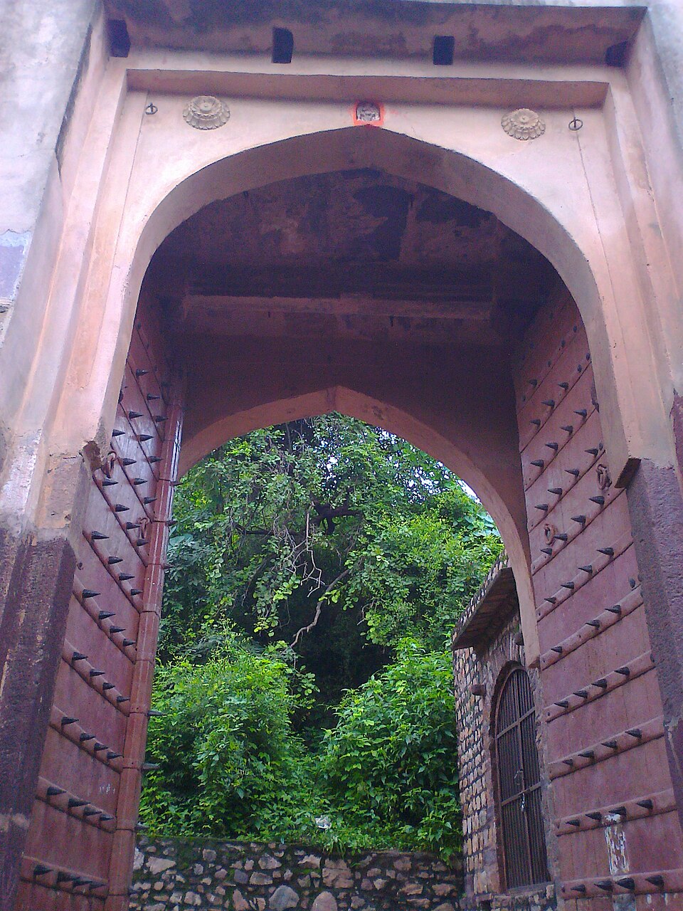 The first gateway of Ranthambore Fort, an arched stone entrance with heavy timber doors studded with iron elephant spikes, opening onto wooded slopes