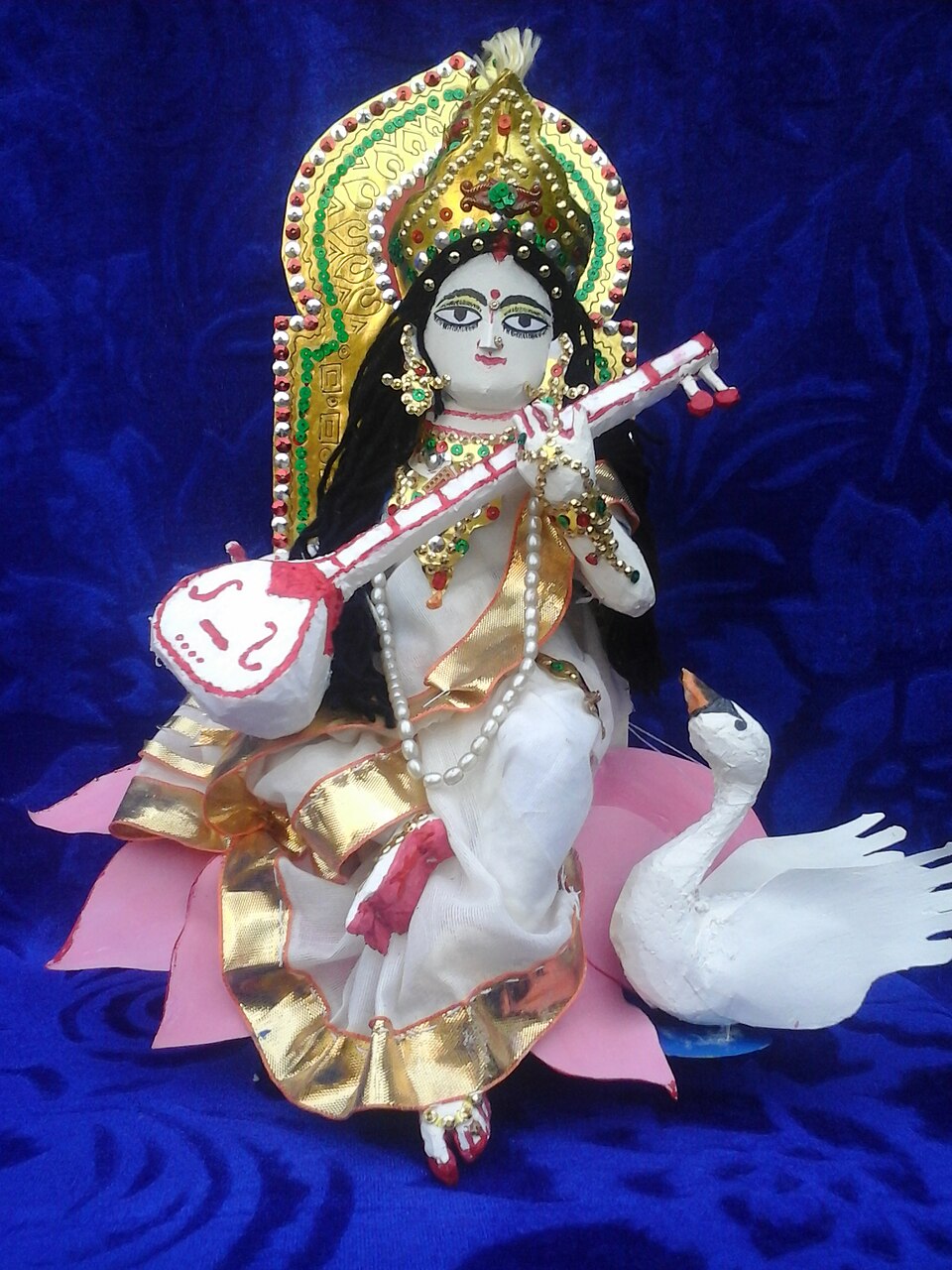 A painted clay image of the goddess Saraswati seated on a lotus in a white sari, holding a veena across her body, with her swan beside her