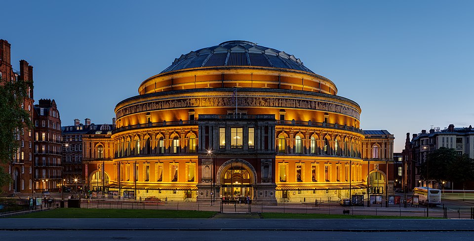 The Royal Albert Hall in London at dusk, its round red-brick and terracotta exterior floodlit beneath the glazed dome