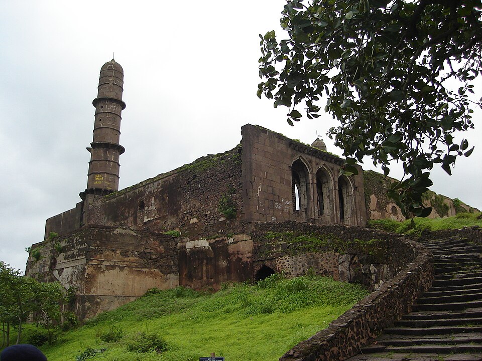 Ruined stone walls and a tall fluted minaret of Asirgarh Fort standing on a grassy hilltop, with steps leading up to the rampart