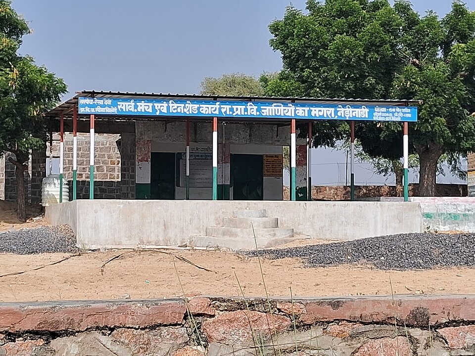 A single-storey rural government primary school building with a tin-shed verandah and a blue Hindi signboard, standing on a concrete plinth in an open yard