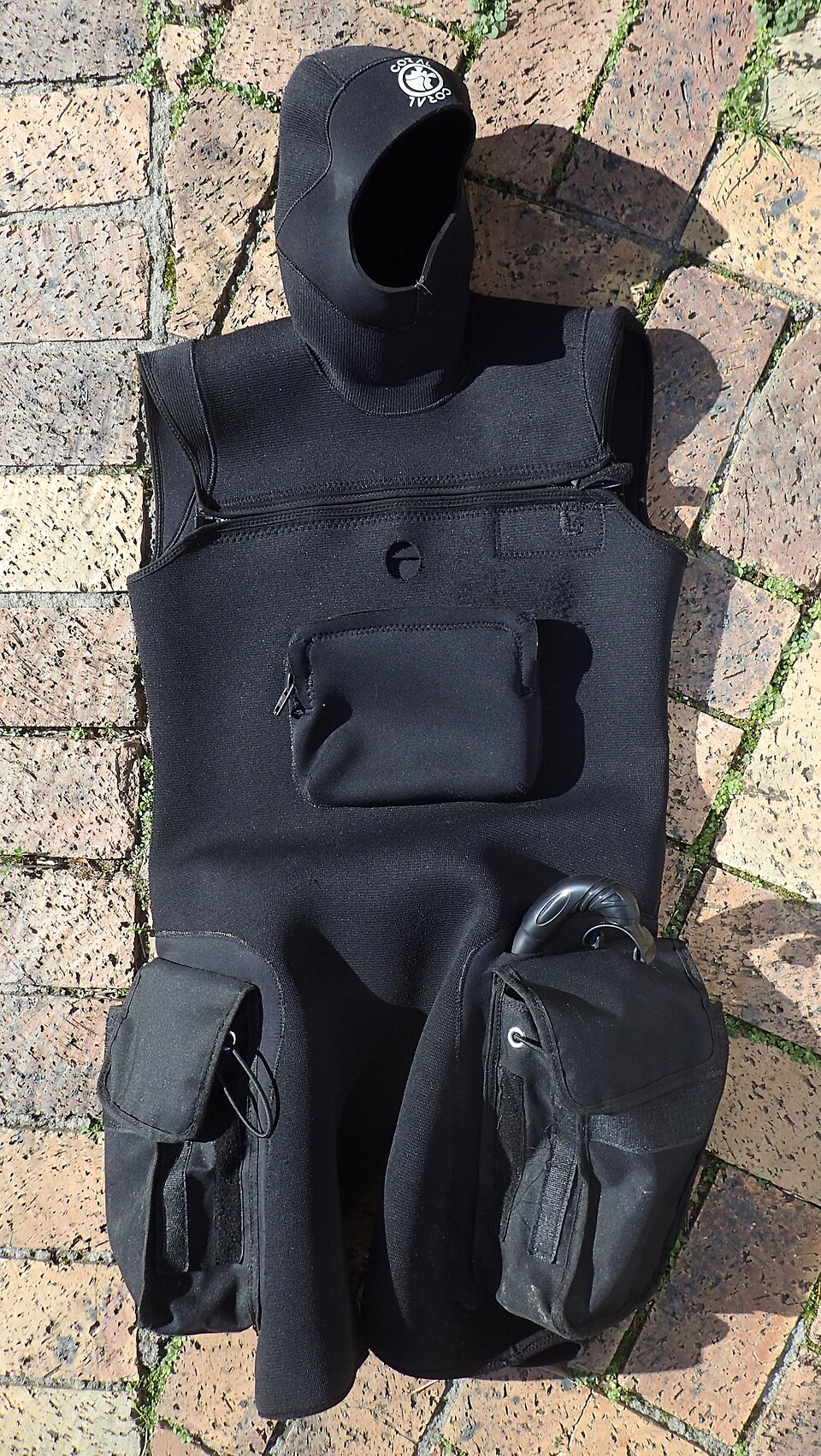 A black neoprene wetsuit tunic with an attached hood and zipped thigh pockets laid flat on paving