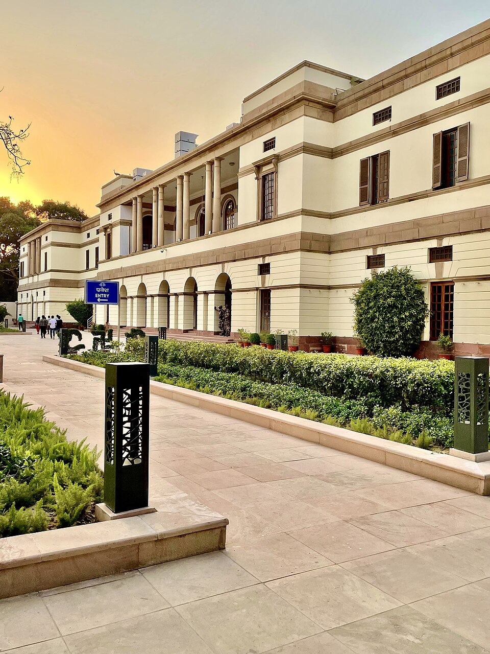 The long colonnaded cream-and-sandstone frontage of Teen Murti Bhavan in New Delhi at sunset, with an entry sign and clipped hedges along the path