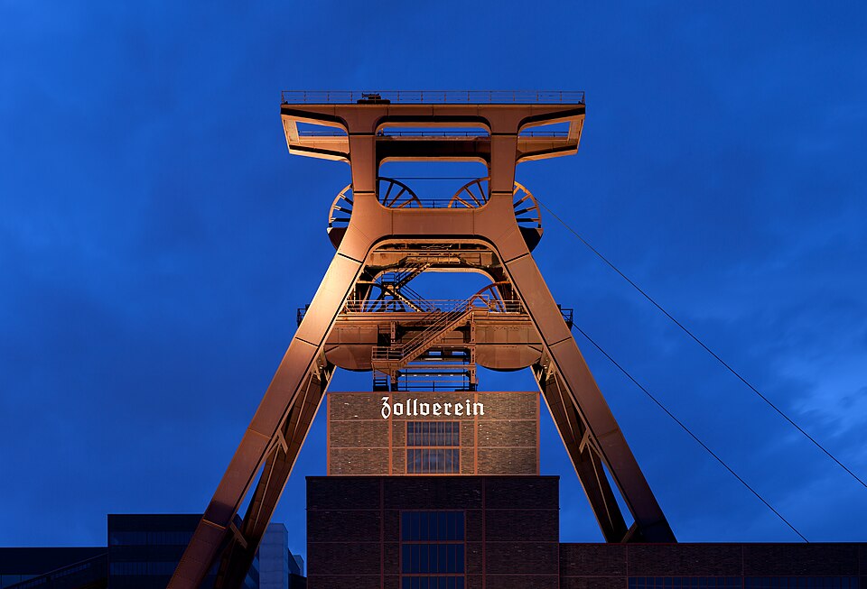 The red double-trestle winding tower of Shaft XII at the Zollverein colliery in Essen, floodlit against a deep blue evening sky above a brick building lettered Zollverein