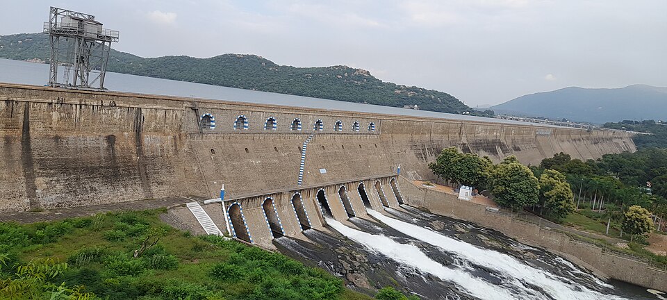 The long masonry wall of Mettur Dam with water pouring from its lower sluices, the reservoir and wooded hills behind
