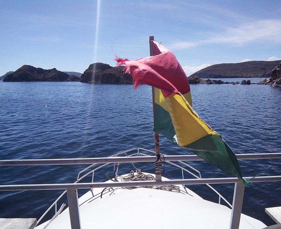 The bow of a boat flying the red, yellow and green Bolivian flag on the deep blue water of Lake Titicaca, with rocky Andean shores behind