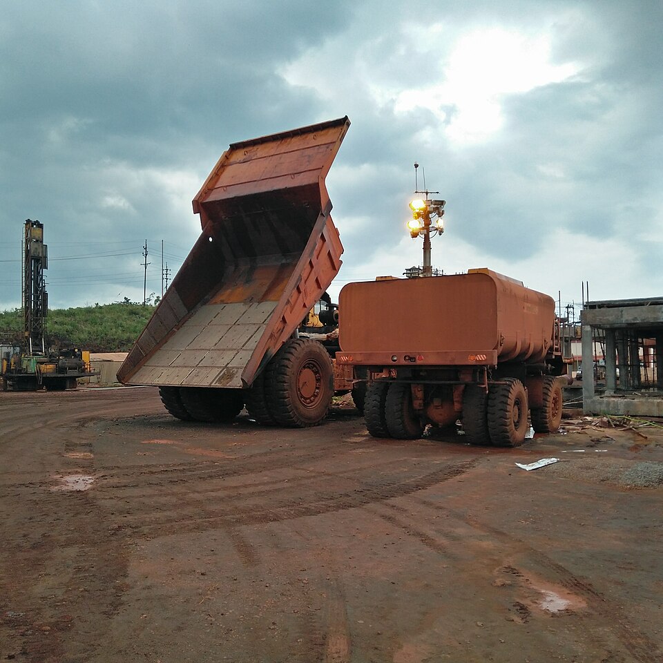Two large haul trucks coated in red iron-ore dust standing on the ore-stained ground of the Bailadila mines, one with its body tipped up