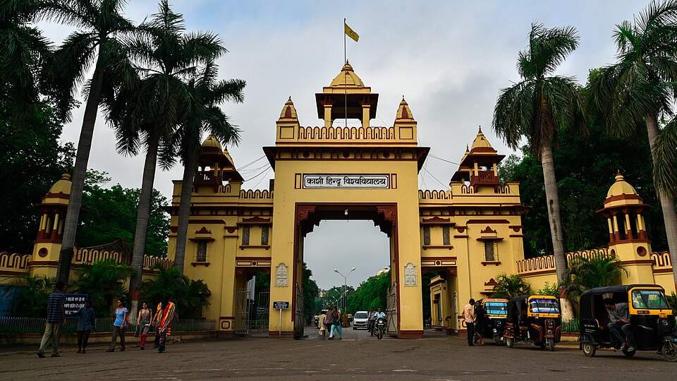 The ochre-coloured main gate of Banaras Hindu University, its arch carrying the name Kashi Hindu Vishwavidyalaya in Devanagari, flanked by palm trees